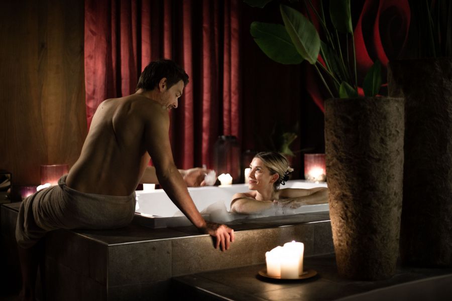 A man sits at the edge of a large tub, a woman covered in foam lies in it, they smile at each other.