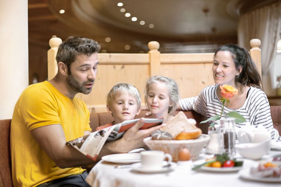 Eine Familie sitzt an einem gedeckten Esstisch, der Vater liest den Kindern aus einer Zeitschrift vor. Eine Familie sitzt an einem gedeckten Esstisch, der Vater liest den Kindern aus einer Zeitschrift vor.