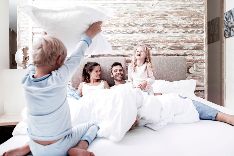 Eine lachende Familie liegt im Doppelbett, die zwei Kinder toben auf dem Bett herum. Eine lachende Familie liegt im Doppelbett, die zwei Kinder toben auf dem Bett herum.